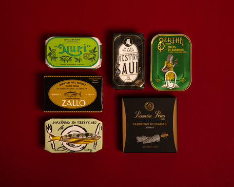 Small selection of tinned fish against a dark red background. One of the tins is bright green and comes from the renowned Portuguese cannery Nuri.