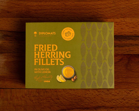 A can of herring in green packaging with yellow lettering against a wood background. There is an image of a small bowl of olive oil with two slices of lemon and two green olives by it on the front of the packaging.