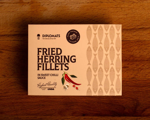 A tin of herring in pale brown card packaging with brown lettering by a picture of two whole red chillies next to two garlic cloves and two slices of red chilli pepper.