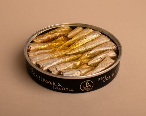 Open tin of small sardines in olive oil. The sardines are very small, you can see around fifteen sardines in the tin. The metal tin is illustrated with the brand name that reads: Real Conservera Espanola 