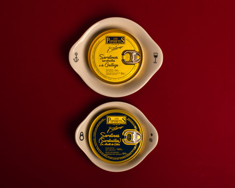 A round tin of sardines with a yellow label on a round eared white ceramic plate with an illustration of an anchor on one ear and a wine glass on the other. This is above another plate with a ring pull on one ear and a shell and heart on the other holding a round tin of sardines with a blue label. The plates rest on a red background. 