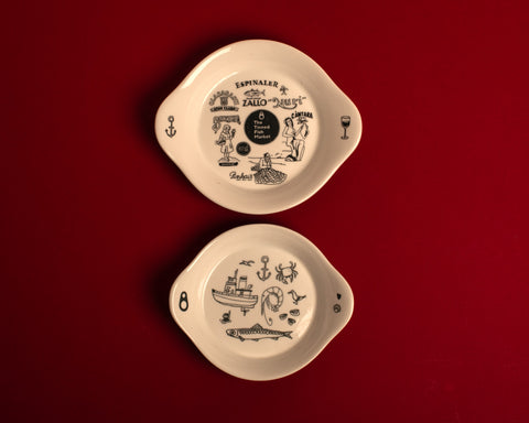 Two illustrated plates, one depicting different brands of Spanish and Portuguese canneries around The Tinned Fish market logo. The one beneath depicting illustrations of a boat, an anchor, a fish, a seagull. The plates are resting on a red background.