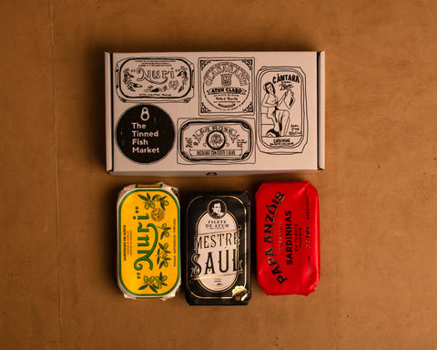 Three colourfully packaged tins of fish beneath a white cardboard gift box illustrated with black line drawings of the tins and The Tinned Fish Market logo.