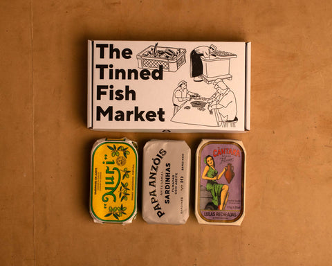 Three colourfully packaged tins of fish beneath a gift box illustrated with line drawings of the cannery workers, a basket of fish and The Tinned Fish Market in bold black lettering.