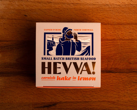 A square box with an image of a fisherman calling out over the Cornish coast. The packaging has a light pink background with purple, orange and gold lettering.