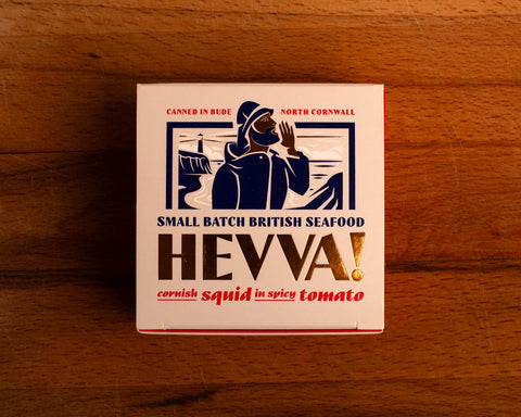 Box containing Cornish squid in spicy tomato from Hevva brand. The box is illustrated with a fisherman fishing on British waters. The box is colourful with the Hevva name in golden letters  