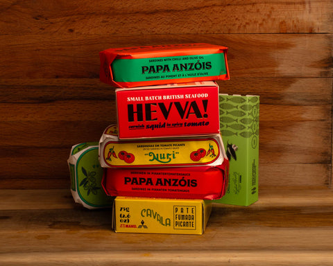 Seven brightly coloured tins of fish some of them illustrated with peppers, against a wood background. All the tins have paper wrappers and all of them are spicy.