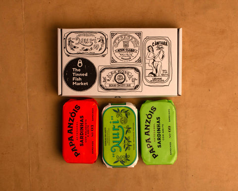 Three colourfully packaged tins of fish beneath a gift box illustrated with line drawings of the tins and The Tinned Fish Market logo.