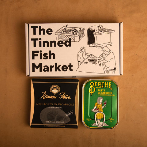 Two tins of fish beneath a gift box illustrated with line drawings of the cannery workers, a basket of fish and The Tinned Fish Market in bold black lettering.