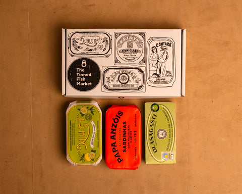 Three colourfully packaged tins of fish beneath a gift box illustrated with line drawings of the tins and The Tinned Fish Market logo.