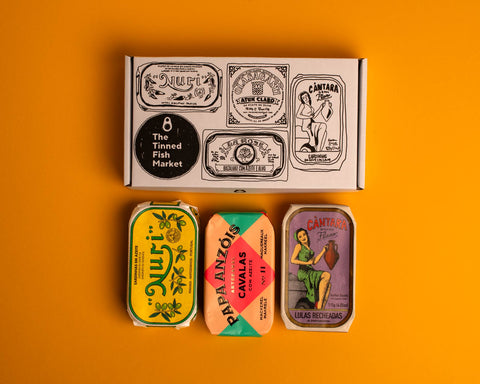 Three colourfully packaged tins of fish beneath a gift box illustrated with line drawings of the tins and The Tinned Fish Market logo.