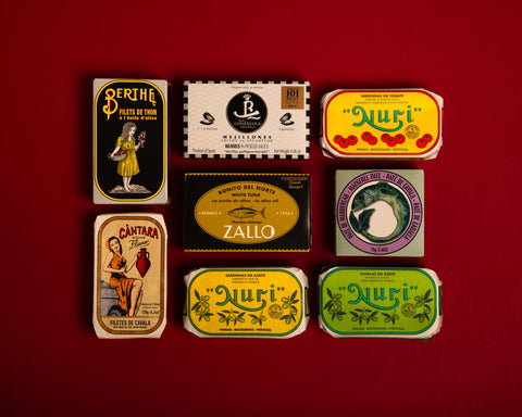 Eight tins on a red table top. The tins have colourful wrappers, one is yellow and is decorated with illustrations  of tomatoes. Another tin has an illustration of a Portuguese lady holding a terracotta vase.   