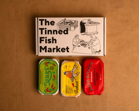 Three colourfully packaged tins of fish beneath a gift box illustrated with line drawings of the cannery workers, a basket of fish and The Tinned Fish Market in bold black lettering.