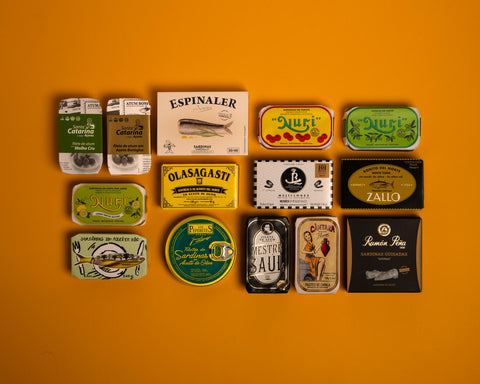Several tins of Spanish and Portuguese tinned fish, two pâtés and a tin of extra spicy sardines arranged on a yellow background.