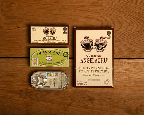 A selection of four small tins and one large of Cantabrian anchovies in classic packaging. The tins are against a wooden backdrop.