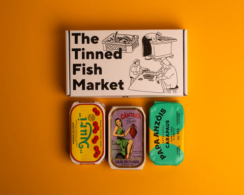 Three colourfully packaged tins of fish beneath a gift box illustrated with line drawings of the cannery workers, a basket of fish and The Tinned Fish Market in bold black lettering.